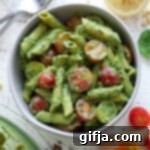 Pesto Pasta Salad 14 Pesto Pasta Salad served in white bowl with tomatoes on wooden board and glass bowl of pasta salad on side. half of lemon and pine nuts and spinach are scattered nearby