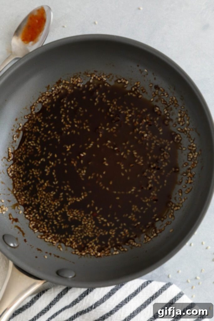 Close-up of the rich, dark brown sesame sauce simmering in a small saucepan, with a spoon resting on the side and a blue and white striped towel nearby, illustrating the sauce preparation.