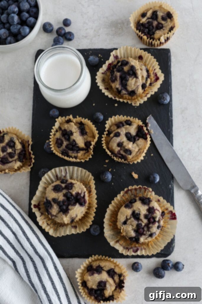 Bakery-Style Vegan Blueberry Muffins 6 Freshly baked Vegan Blueberry Muffins served on a black rectangular platter with a glass of almond milk, a knife, and scattered fresh blueberries.