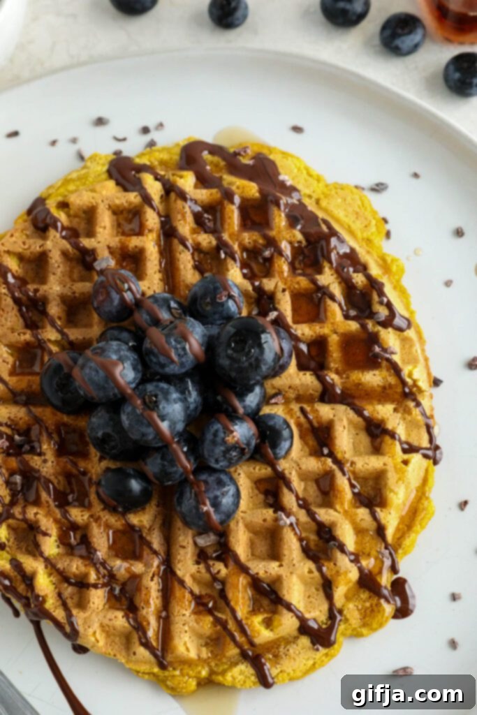 Three golden Pumpkin Turmeric Waffles topped with fresh blueberries, crunchy cacao nibs, and a rich chocolate drizzle, served on a pristine white plate