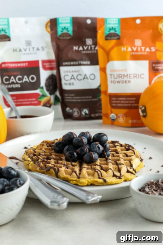 A stack of freshly made Pumpkin Turmeric Waffles, topped with blueberries, cacao nibs, and a decadent chocolate drizzle, showcased on a white plate